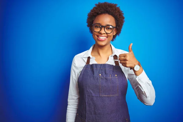 Young african american woman shop owner wearing business apron over blue background doing happy thumbs up gesture with hand. Approving expression looking at the camera with showing success.