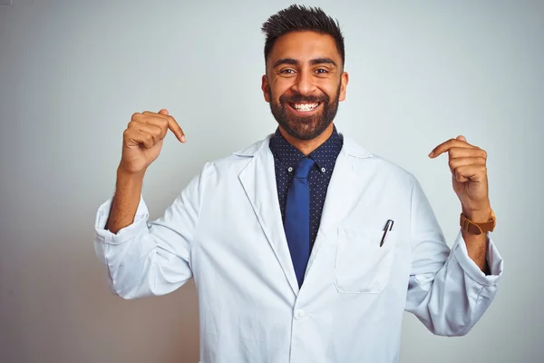 Young indian doctor man standing over isolated white background looking confident with smile on face, pointing oneself with fingers proud and happy.