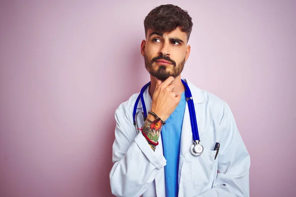 Young doctor man with tattoo wearing stethocope standing over isolated pink background with hand on chin thinking about question, pensive expression. Smiling with thoughtful face. Doubt concept.