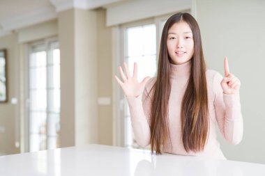 Güzel Asyalı kadın beyaz masada rahat bir süveter giyer ve altı numaralı parmaklarıyla işaret ederken kendinden emin ve mutlu bir şekilde gülümser..