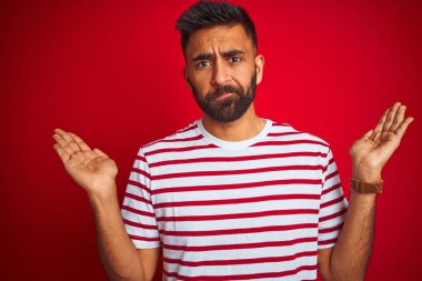 İzole kırmızı arka plan üzerinde duran çizgili t-shirt giyen genç Hintli adam habersiz ve kolları ve elleri ile karışık ifade kaldırdı. Şüphe kavramı.