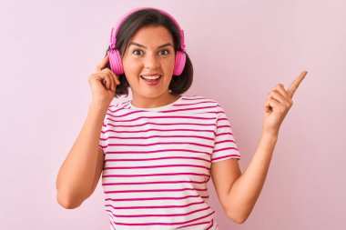Genç ve güzel bir kadın müzik dinliyor. Pembe arka planda kulaklık takıyor. Elleri ve parmakları kenarı işaret ederek çok mutlu.