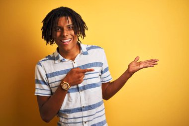 Izole sarı arka plan üzerinde çizgili gömlek giyen dreadlocks ile Afro Amerikan adam şaşırttı ve el ile sunarken kameraya gülümseyerek ve parmak ile işaret.