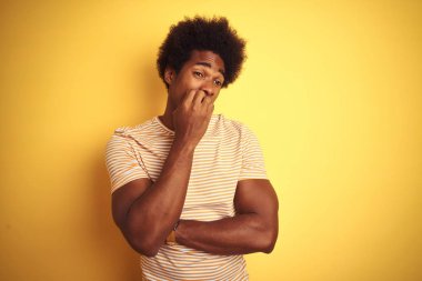 Afro saçlı Amerikalı adam çizgili t-shirt izole sarı arka plan üzerinde duran giyen stresli ve ağız tırnak ısırma elleri ile sinir görünümlü. Anksiyete sorunu.