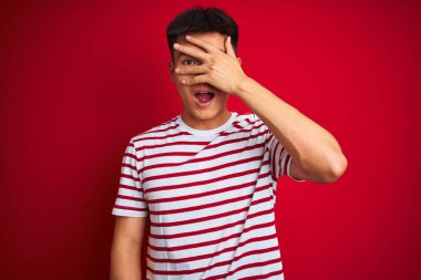 İzole kırmızı arka plan üzerinde duran çizgili t-shirt giyen genç Asyalı çinli adam şok el ile yüz ve gözleri kapsayan gözetleme, utanmış ifade ile parmaklarından bakarak.