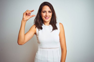 İzole edilmiş beyaz arka planda duran elbiseler giyen genç ve güzel bir kadın gülümsüyor ve kendinden emin bir şekilde el hareketi yapıyor parmaklarıyla işaret yapıyor ve kameraya bakıyor. Ölçü kavramı.