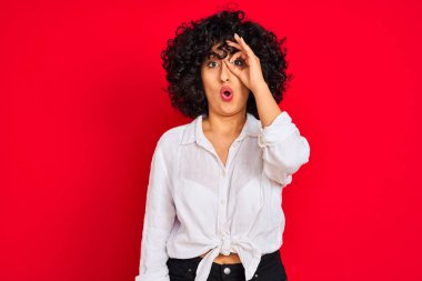 Kıvırcık saçlı genç arap kadın izole kırmızı arka plan üzerinde beyaz rahat gömlek giyen ok jest yaparken şaşırmış yüz ile şok, parmaklardan bakarak göz. İnanmayan ifade.