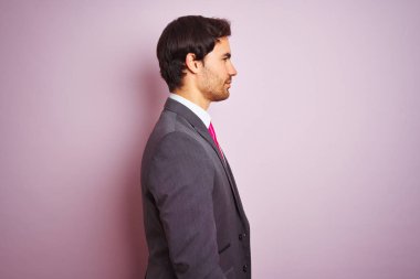 Takım elbise giyen ve kravat giyen genç yakışıklı işadamı yan bakarak izole pembe arka plan üzerinde duran, kendine güvenen gülümseme ile doğal yüzü ile profil poz rahatlayın.