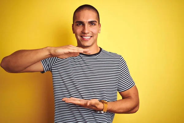 年轻英俊的男子穿着条纹T恤 头戴黄色的孤立背景图 手握大而大的标志 尺寸符号 笑着看着摄像机 计量概念 — 图库照片