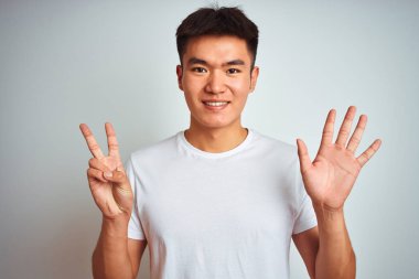 Genç asyalı Çinli adam izole beyaz arka plan üzerinde duran t-shirt giyen gösteren ve parmakları ile işaret ederken emin ve mutlu gülümseyerek yedi numara.