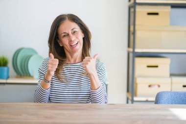 Orta yaş yaşlı kadın evde masada oturan başarı işareti el ile olumlu jest yapıyor, gülümseyerek ve mutlu başparmak kadar. Neşeli ifade ve kazanan jest.