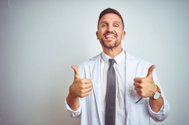 Genç yakışıklı doktor adam izole arka plan başarı işareti üzerinde beyaz profressional ceket giyen el ile olumlu jest yapıyor, gülümseyen ve mutlu başparmak kadar. Neşeli ifade ve kazanan jest.