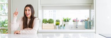 Evde beyaz masada oturan güzel genç kadının geniş açılı resmi başarılı bir fikirle parmağını yukarı doğru gösteriyor. Çıktı ve mutlu. Bir numara.