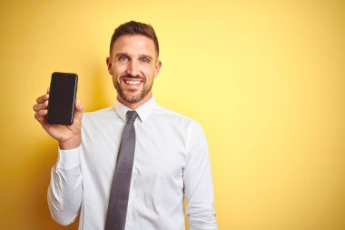 Genç yakışıklı iş adamı ayakta mutlu bir yüz ve dişleri gösteren kendinden emin bir gülümseme ile gülümseyen sarı izole arka plan üzerinde akıllı telefon ekranı gösteren
