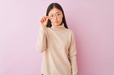 Genç çinli kadın ciddi düşünme akıllı yüz üzerinde kendine güvenen bir ifade ile izole pembe arka plan üzerinde balıkçı kazak ve gözlük giyiyor