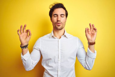 Genç yakışıklı işadamı izole sarı arka plan üzerinde duran zarif gömlek giyen dinlenmek ve gözleri parmakları ile meditasyon jest yapıyor kapalı gülümseyerek. Yoga konsepti.
