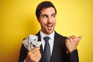 İzole sarı arka plan üzerinde dolar tutan takım elbise giyen genç yakışıklı işadamı işaret ve mutlu yüzü gülümseyerek yan başparmak ile gösteren