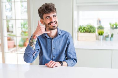 Beyaz masanın üzerinde oturan rahat gömlek giyen genç adam telefonda konuşmak gibi el ve parmaklarıyla telefon hareketi yapıyor gülümseyerek. İletişim kavramları.