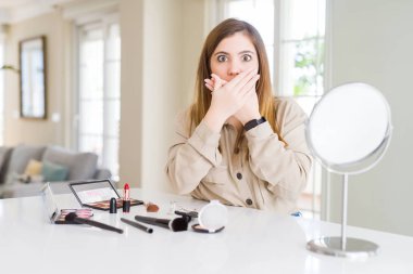 Beautiful young woman using make up cosmetics shocked covering mouth with hands for mistake. Secret concept.