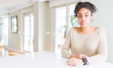 Güzel genç afro-amerikan kadın afro saçlı evde komik yüzü ile yanakları şişirme masada oturan. Ağız hava ile şişirilmiş, deli ifade.