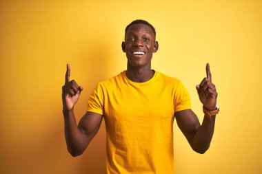 Genç afro-Amerikan adam izole sarı arka plan üzerinde duran rahat t-shirt giyen şaşırmış ve şaşırttı ve parmakları ile işaret ve kolları kaldırdı.