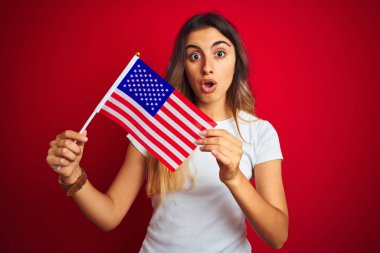 Amerika Birleşik Devletleri bayrağını kırmızı izole arka plan üzerinde tutan genç kadın sürpriz bir yüzle şokta korkmuş, korku ve korku ifadesi ile heyecanlı