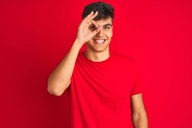 İzole edilmiş kırmızı arka plan üzerinde duran genç Hintli adam el hareketleriyle gülümsüyor ve mutlu yüzle parmaklarının arasından bakıyor..