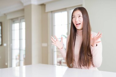 Güzel bir Asyalı kadın beyaz masada rahat bir kazak giyiyor deli gibi bağırıyor ve agresif bir ifadeyle bağırıyor ve kollarını kaldırıyor. Hayal kırıklığı konsepti.