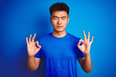 İzole mavi arka plan üzerinde duran t-shirt giyen genç Asyalı Çinli adam rahatlayın ve parmakları ile meditasyon jest yapıyor kapalı gözleri ile gülümseyerek. Yoga konsepti.