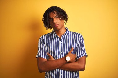 Izole sarı arka plan şüpheci ve sinir üzerinde rahat çizgili t-shirt giyen dreadlocks ile Afro adam, çapraz kolları ile yüzünde onaylamayan ifade. Negatif kişi.