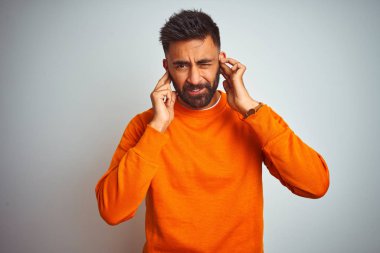 Genç hintli adam yüksek sesle müzik sesi için rahatsız ifade ile parmakları ile kulakları kapsayan izole beyaz arka plan üzerinde turuncu kazak giyiyor. Sağır kavramı.