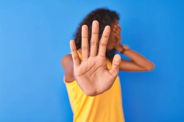 Young brazilian woman wearing yellow t-shirt standing over isolated blue background covering eyes with hands and doing stop gesture with sad and fear expression. Embarrassed and negative concept.