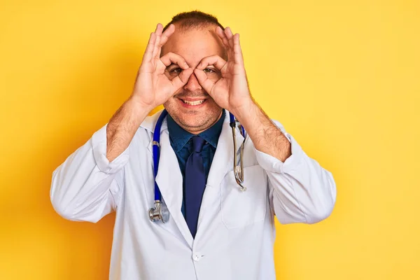 Young doctor man wearing coat and stethoscope standing over isolated yellow background doing ok gesture like binoculars sticking tongue out, eyes looking through fingers. Crazy expression.