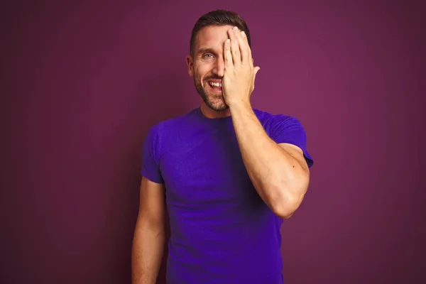 Young man wearing casual purple t-shirt over lilac isolated background covering one eye with hand, confident smile on face and surprise emotion.