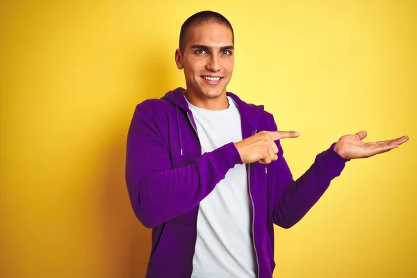 年轻英俊的男子穿着紫色运动衫 披着黄色孤立的背景 一边手牵着手 一边指指点点着相机 一边惊讶地微笑着 — 图库照片