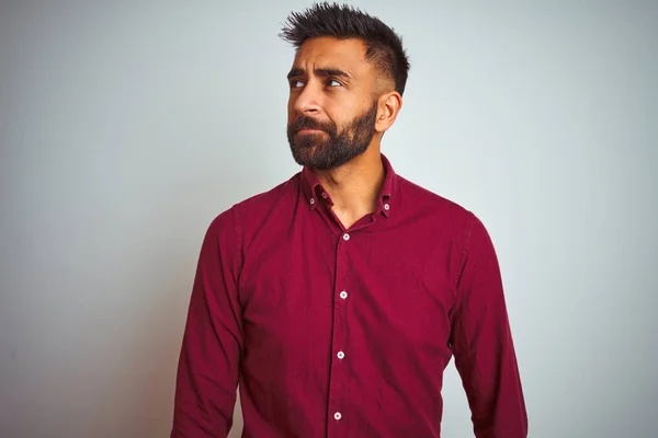 Young indian man wearing red elegant shirt standing over isolated grey background smiling looking to the side and staring away thinking.