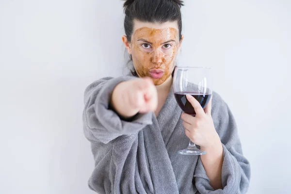 Beautiful woman wearing cosmetic facial mask as skincare treatment drinking glass of wine pointing with finger to the camera and to you, hand sign, positive and confident gesture from the front