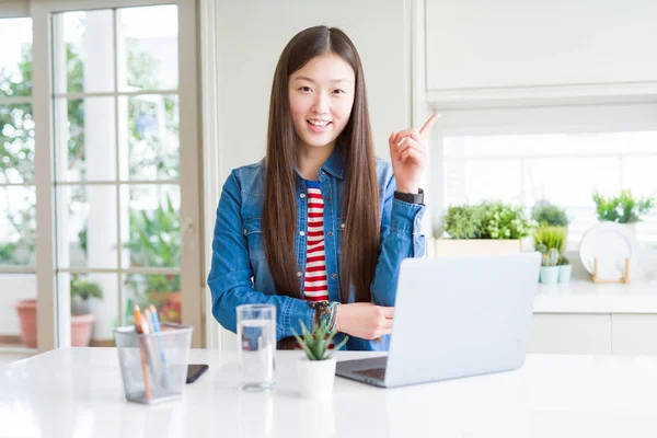 Beautiful Asian woman working using computer laptop with a big smile on face, pointing with hand and finger to the side looking at the camera.