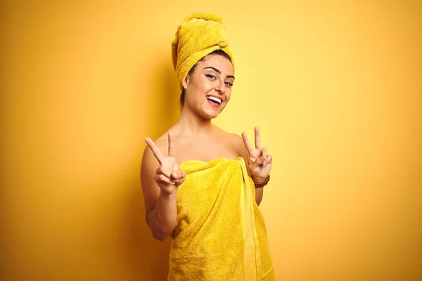 Young beautiful woman wearing towel after shower over isolated yellow background smiling looking to the camera showing fingers doing victory sign. Number two.