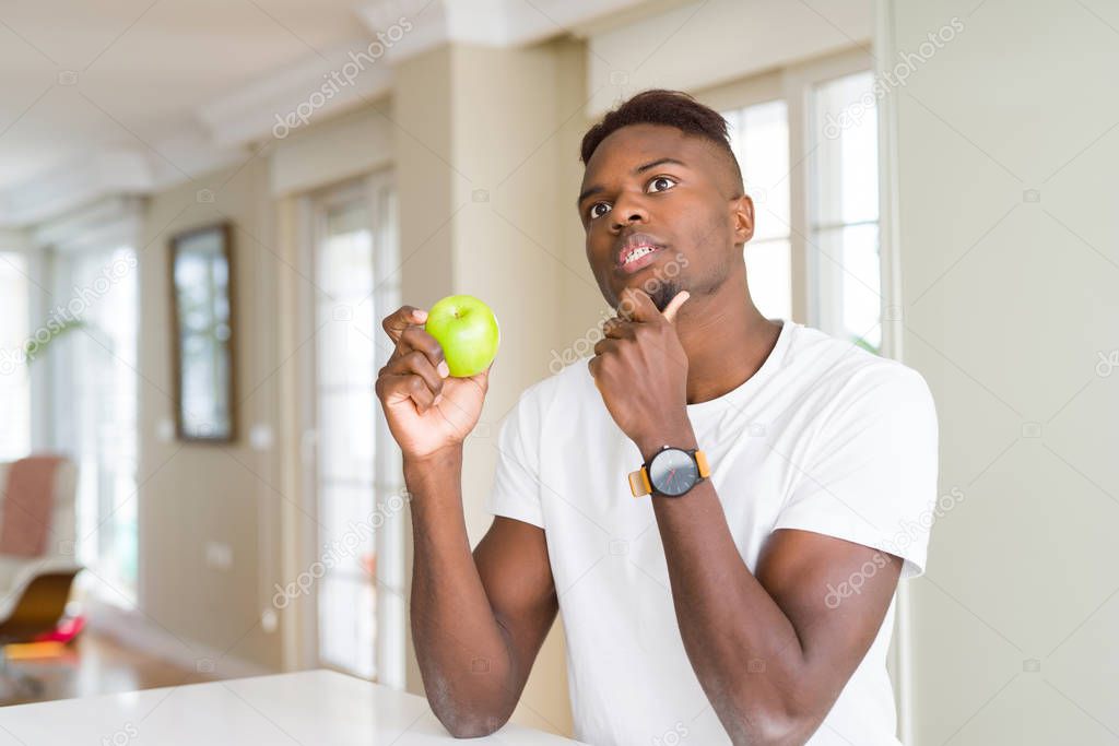 Young african american man eating fresh green apple serious face thinking about question, very confused idea