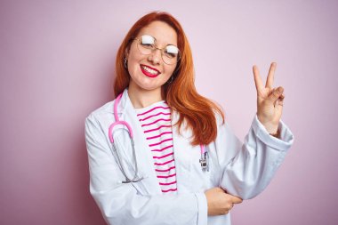 Pembe izole edilmiş arka planda gözlük takan genç kızıl saçlı kadın kameraya gülümseyen ve zafer işareti yapan. İki numara..