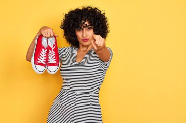 Kıvırcık saçlı genç arap kadın kamera ve size parmak ile işaret izole sarı arka plan üzerinde rahat spor ayakkabı tutan, el işareti, önden olumlu ve kendinden emin jest