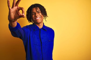 Afro worker man with dreadlocks wearing mechanic uniform over isolated yellow background smiling positive doing ok sign with hand and fingers. Successful expression.