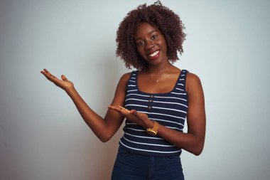 İzole beyaz arka plan üzerinde duran çizgili t-shirt giyen genç Afrikalı afro kadın şaşırttı ve el ile sunarken kameraya gülümseyerek ve parmak ile işaret.