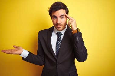 Takım elbise giyen ve izole sarı arka plan üzerinde duran kravat giyen genç yakışıklı işadamı şaşkın ve açık palmiye ile rahatsız kopya alanı gösteren ve alnına parmak işaret. Bir düşün..