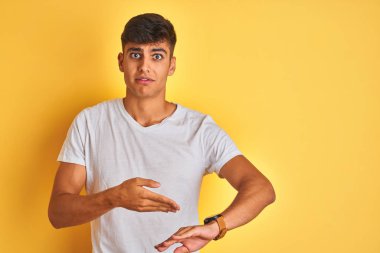 İzole edilmiş sarı arka plan üzerinde duran beyaz t-shirt giyen genç Hintli adam saati, sabırsızlığı ve gecikmeyi izlemek için acele ediyor.