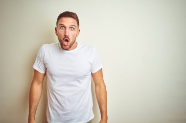 İzole edilmiş arka plan üzerinde gündelik beyaz t-shirt giyen genç yakışıklı adam şaşkınlık ve şaşkınlık dolu bir ifade, korku ve heyecanlı bir yüz ifadesiyle şoka uğradı..
