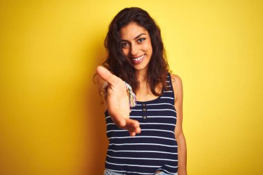 İzole sarı arka plan üzerinde duran çizgili t-shirt giyen genç güzel kadın yardım ve kabul veren palmiye el sunan neşeli gülümseyerek.