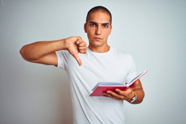 Genç yakışıklı adam, beyaz izole edilmiş arka plan üzerinde kırmızı kitap okuyor. Kızgın yüz ifadesi, olumsuz işaret baş parmakları aşağıda, reddedilme konsepti ise olumsuz.