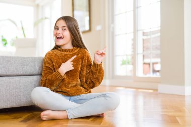 Güzel genç kız çocuk evde katta oturan gülümseyen ve iki elleri ve parmakları ile tarafına işaret kamera bakıyor.
